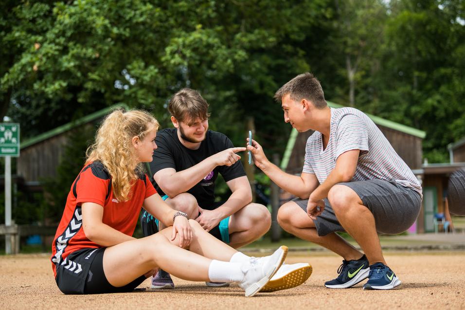 Drei Jugendliche sitzen auf einem Sandplatz und betrachten gemeinsam ein Smartphone. Bäume und Hütten im Hintergrund.