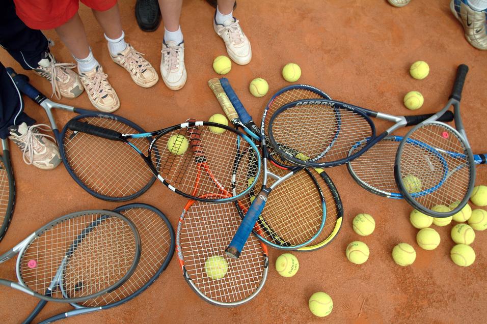 Ansammlung von Tennisschlägern und Bällen auf einem Sandplatz, umgeben von Füßen in Sportschuhen.