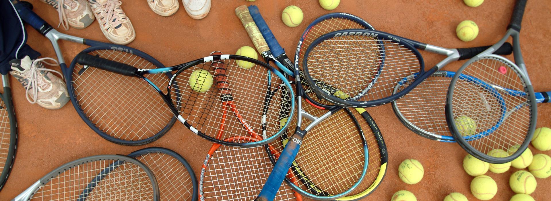 Ansammlung von Tennisschlägern und Bällen auf einem Sandplatz, umgeben von Füßen in Sportschuhen.