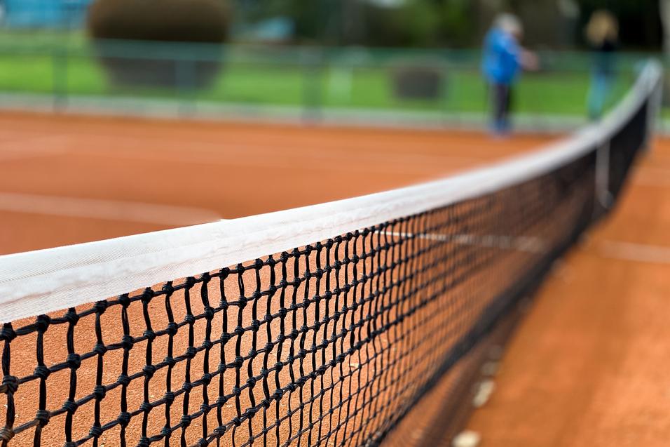 Nahaufnahme eines Tennisnetzes auf einem roten Sandplatz, Hintergrund unscharf mit grünen Bäumen.