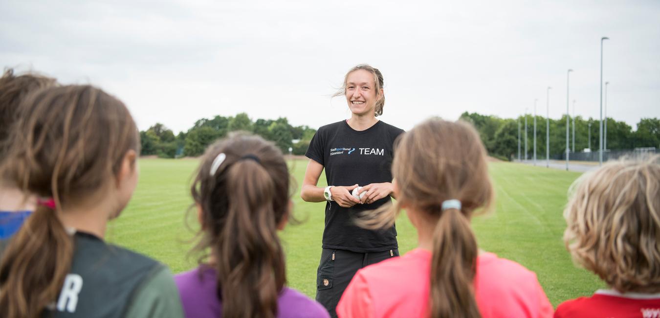 Trainerin spricht mit jungen Sportlerinnen auf einem grünen Fußballfeld unter bewölkten Himmel.